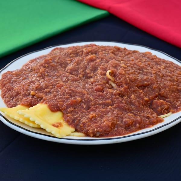 White plate of ravioli and spaghetti covered in a chunky red sauce, with green and red fabric behind.