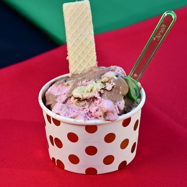 Multi-flavored ice cream in a polka-dotted cup with a wafer and green spoon on a colorful background.